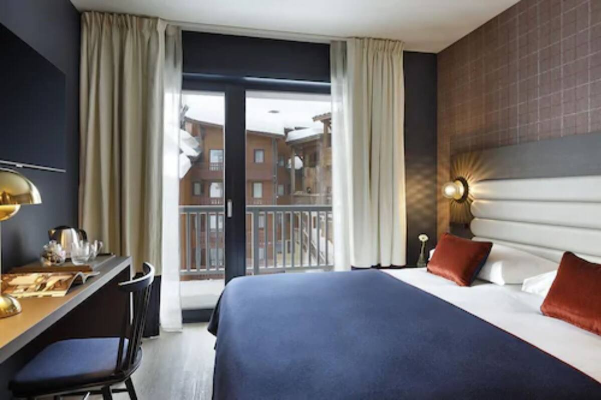 Habitación de hotel con una cama grande, un escritorio con lámpara y vistas a edificios a través del ventanal.