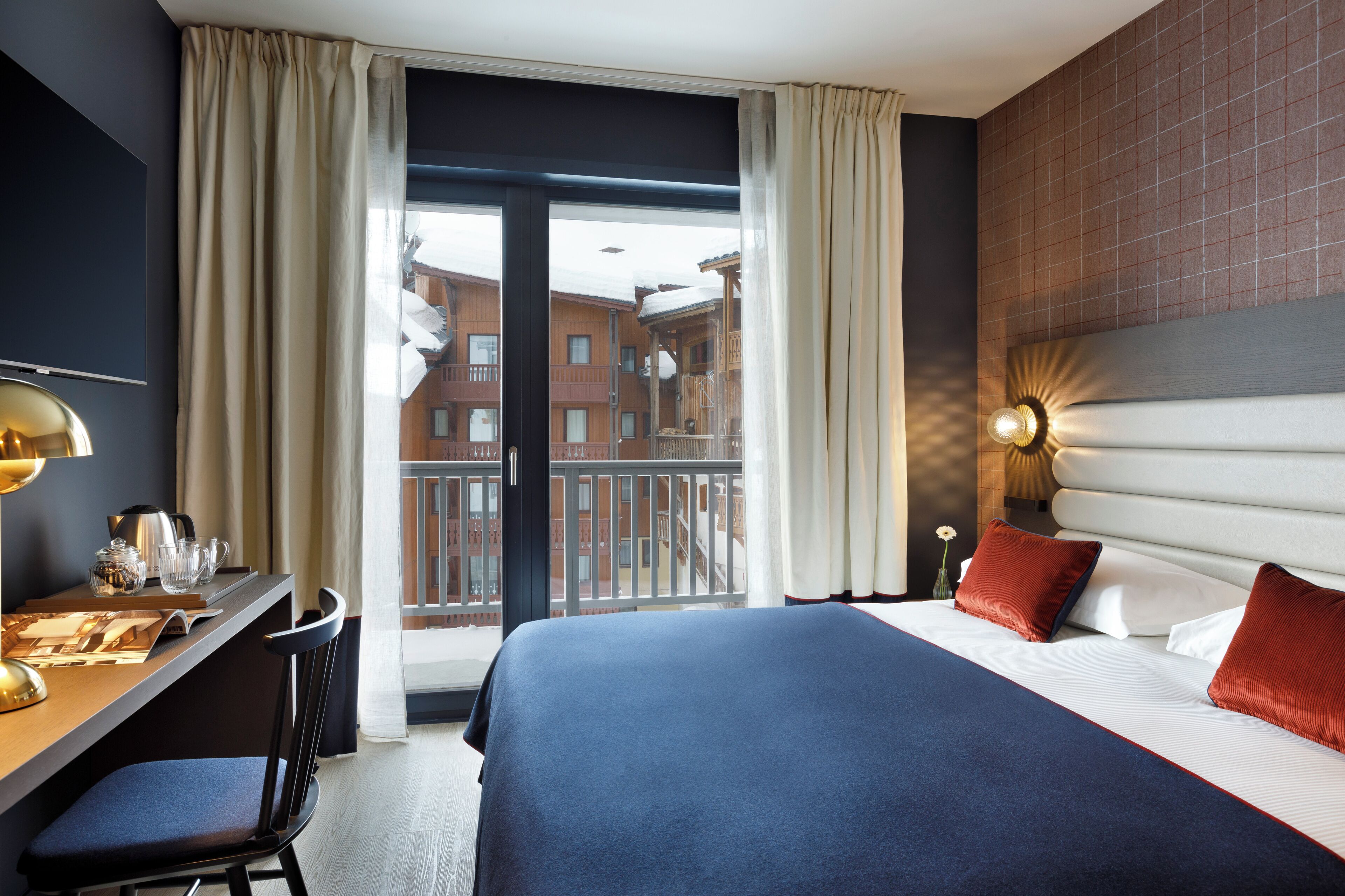 Hotelkamer met een bed, een bureau met stoel, een televisie en een balkon met uitzicht op gebouwen.