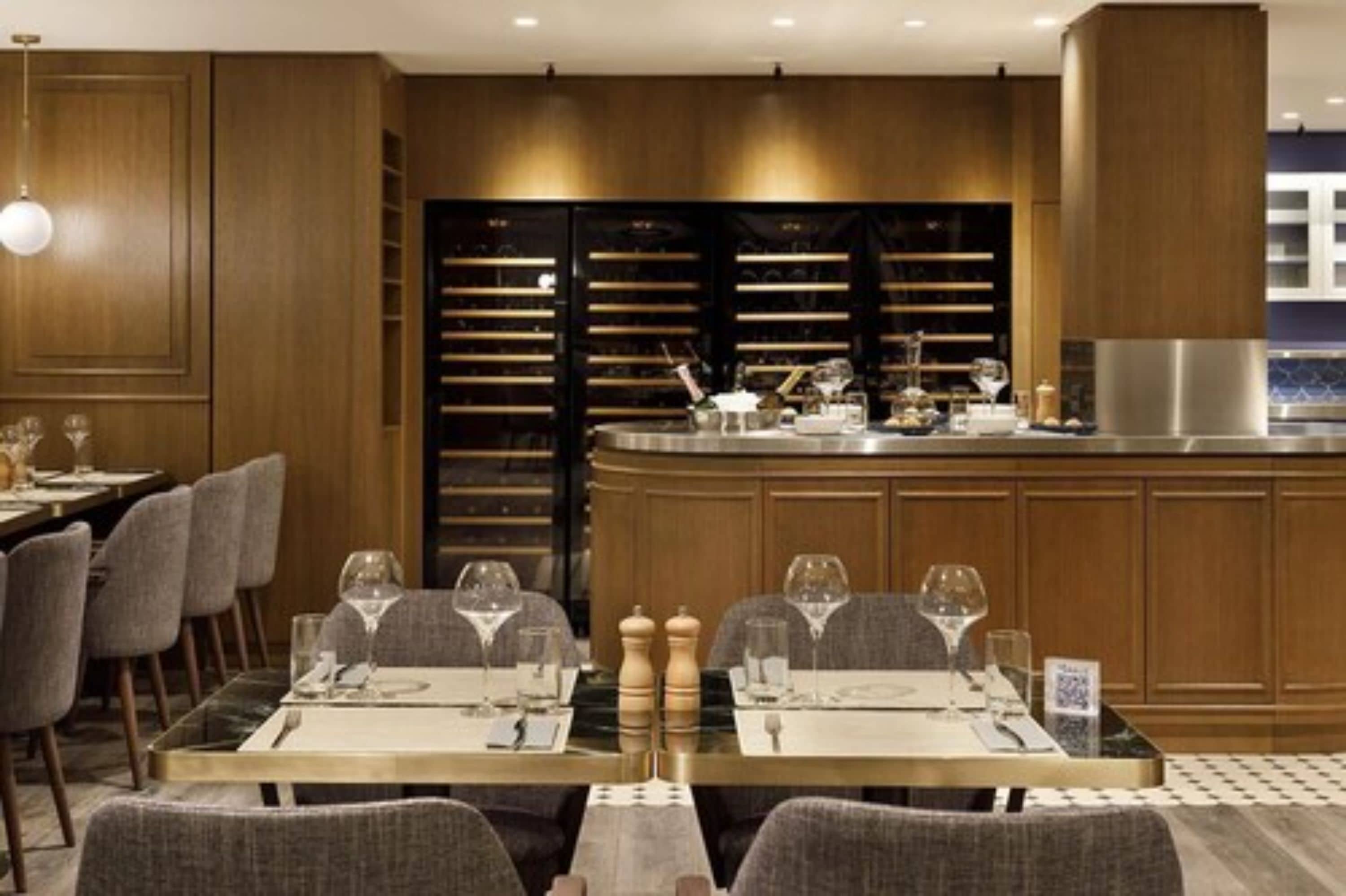Un restaurant moderne doté de meubles en bois, d’un îlot central avec des ustensiles de bar et d’un espace repas avec des tables prévues pour deux personnes.