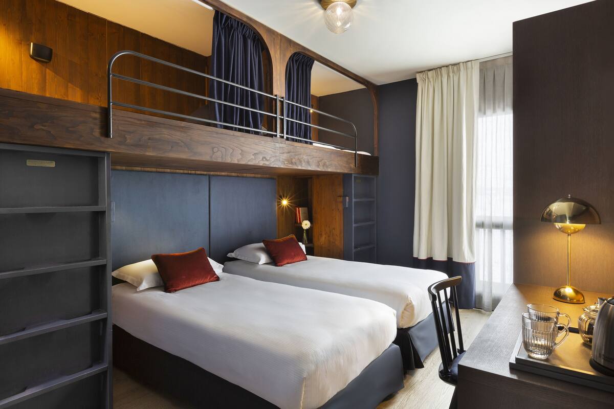 Habitación de hotel con dos camas, un escritorio, una silla, una lámpara y una ventana con cortinas.