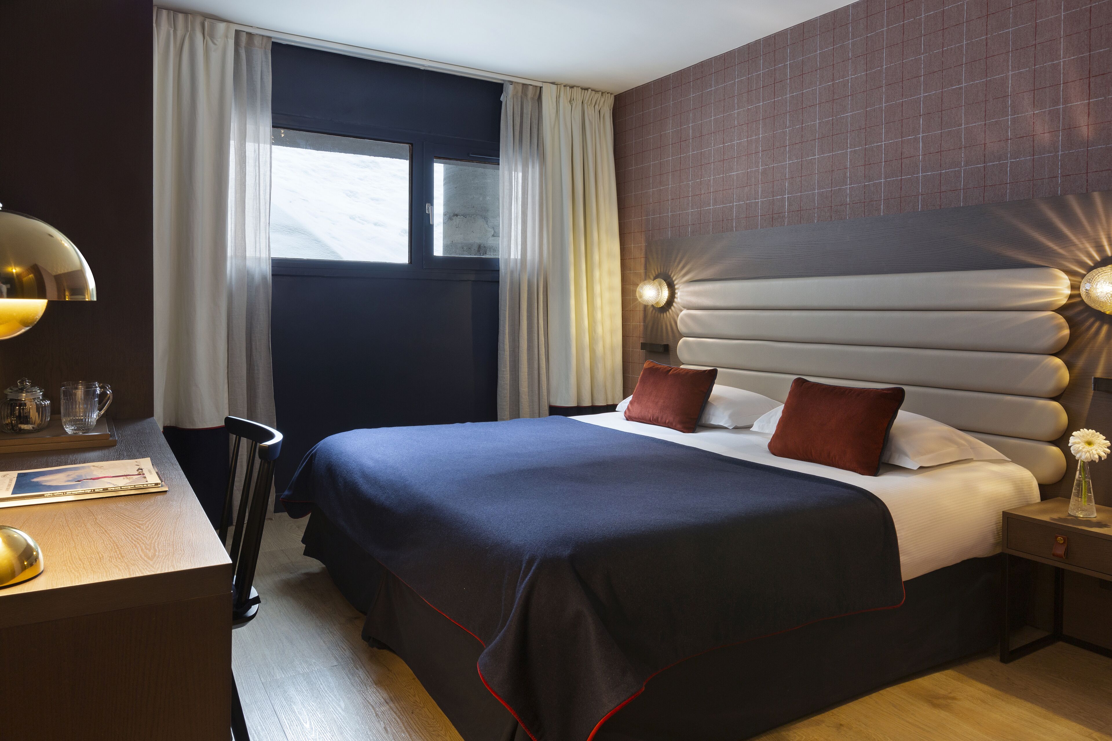 A hotel room with a large bed, a desk with a lamp, a window with curtains, and a wall with a textured pattern.
