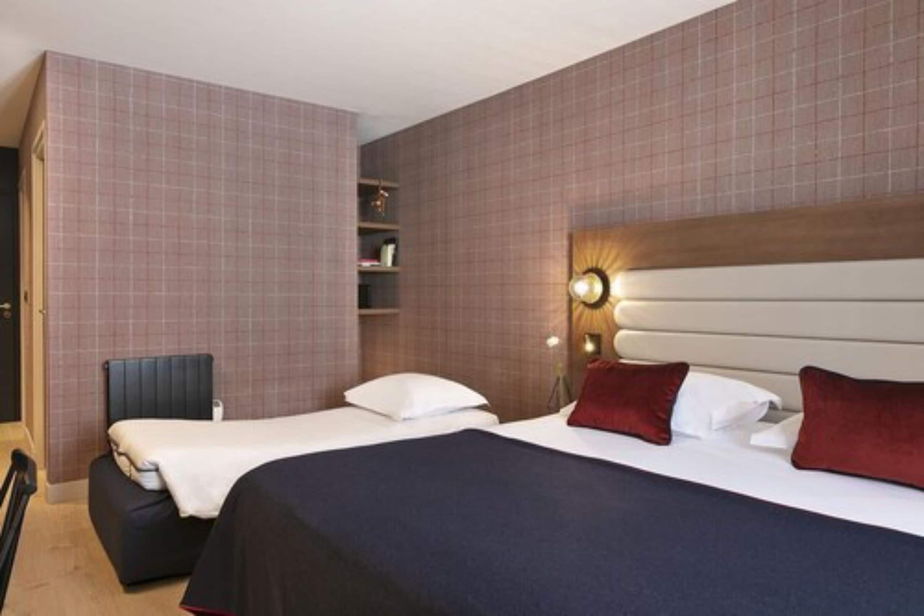 Habitación de hotel con dos camas, cabecera iluminada, un pequeño rincón de descanso y una pared con diseño de cuadrícula.
