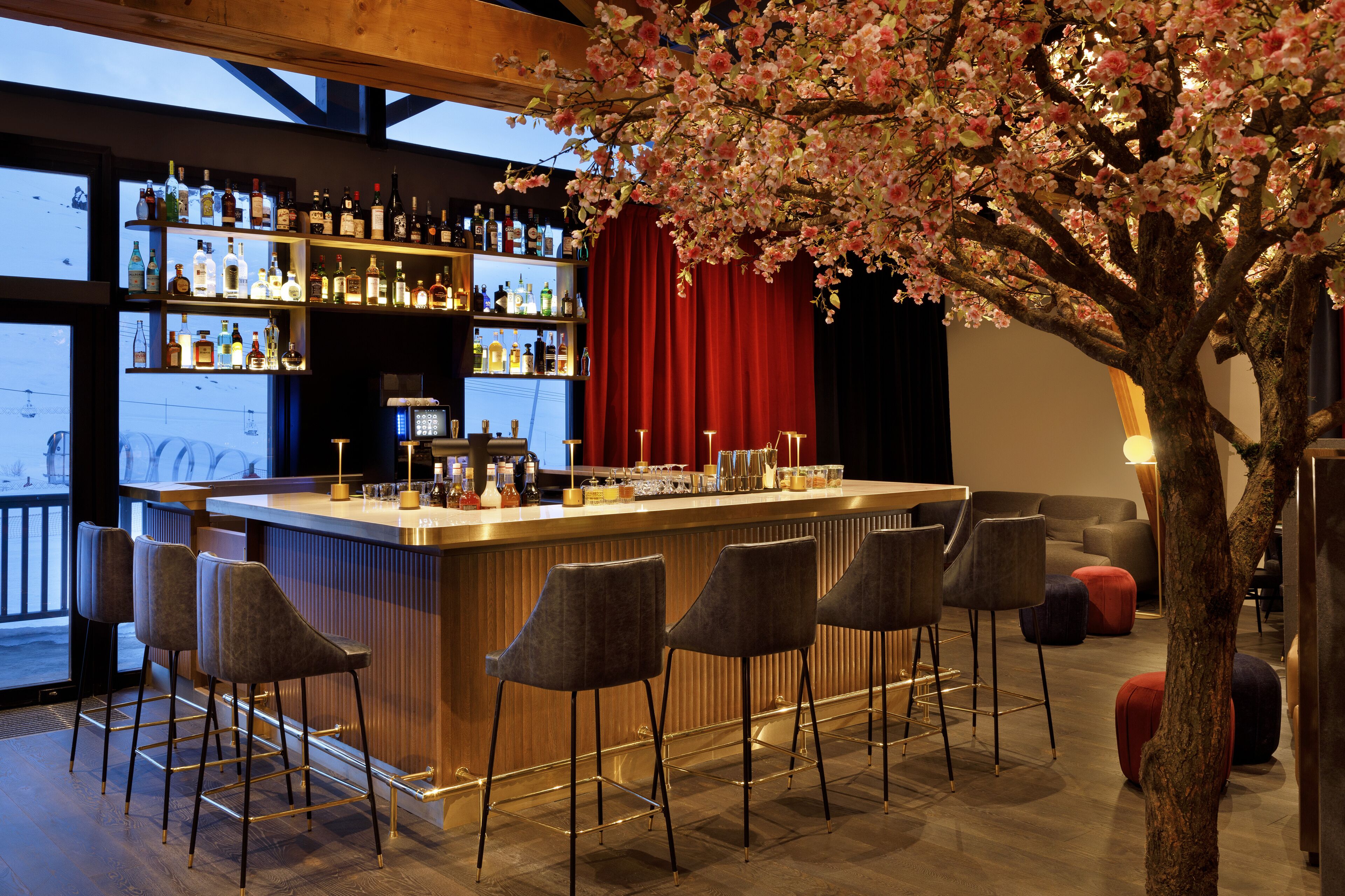 A bar area with a wooden bar counter, multiple stools, and a large tree with pink blossoms. Shelves with bottles are visible in the background.