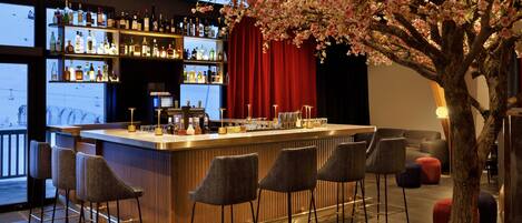 A bar area with a wooden bar counter, multiple stools, and a large tree with pink blossoms. Shelves with bottles are visible in the background.