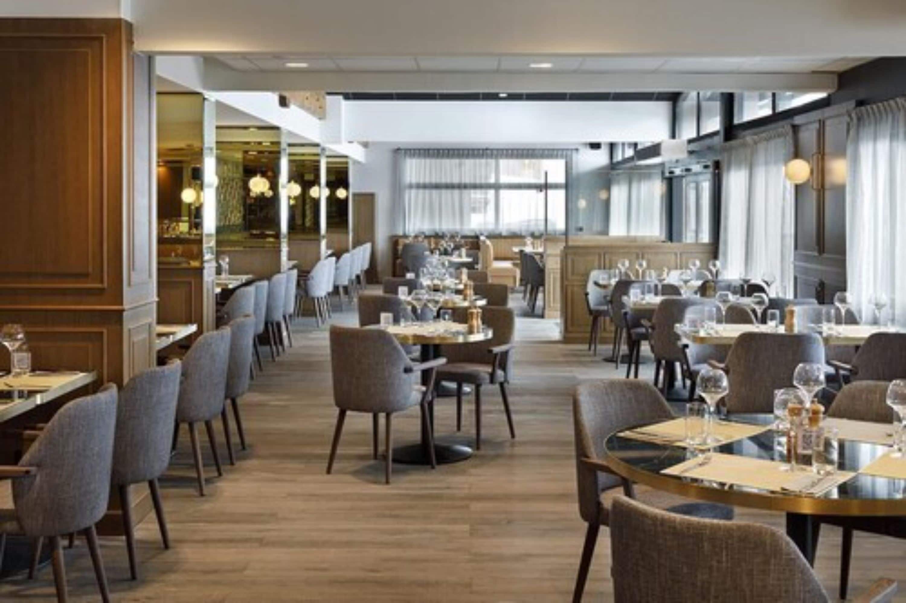 A restaurant with multiple dining tables and chairs, a wooden floor, and large windows with curtains.
