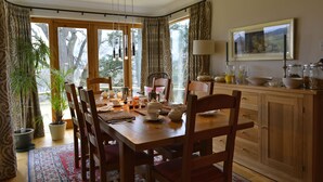 A dining area with a wooden table, chairs, and a view through large windows. - Brae House Bed & Breakfast (Aberfeldy)
