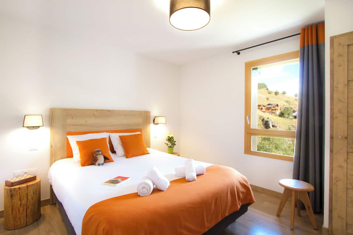 A neatly made bed with orange bedding, a wooden headboard, and a view of a rural landscape through a window.