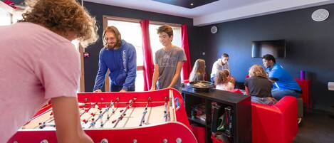 A room with a foosball table, red seating, and a television on the wall.