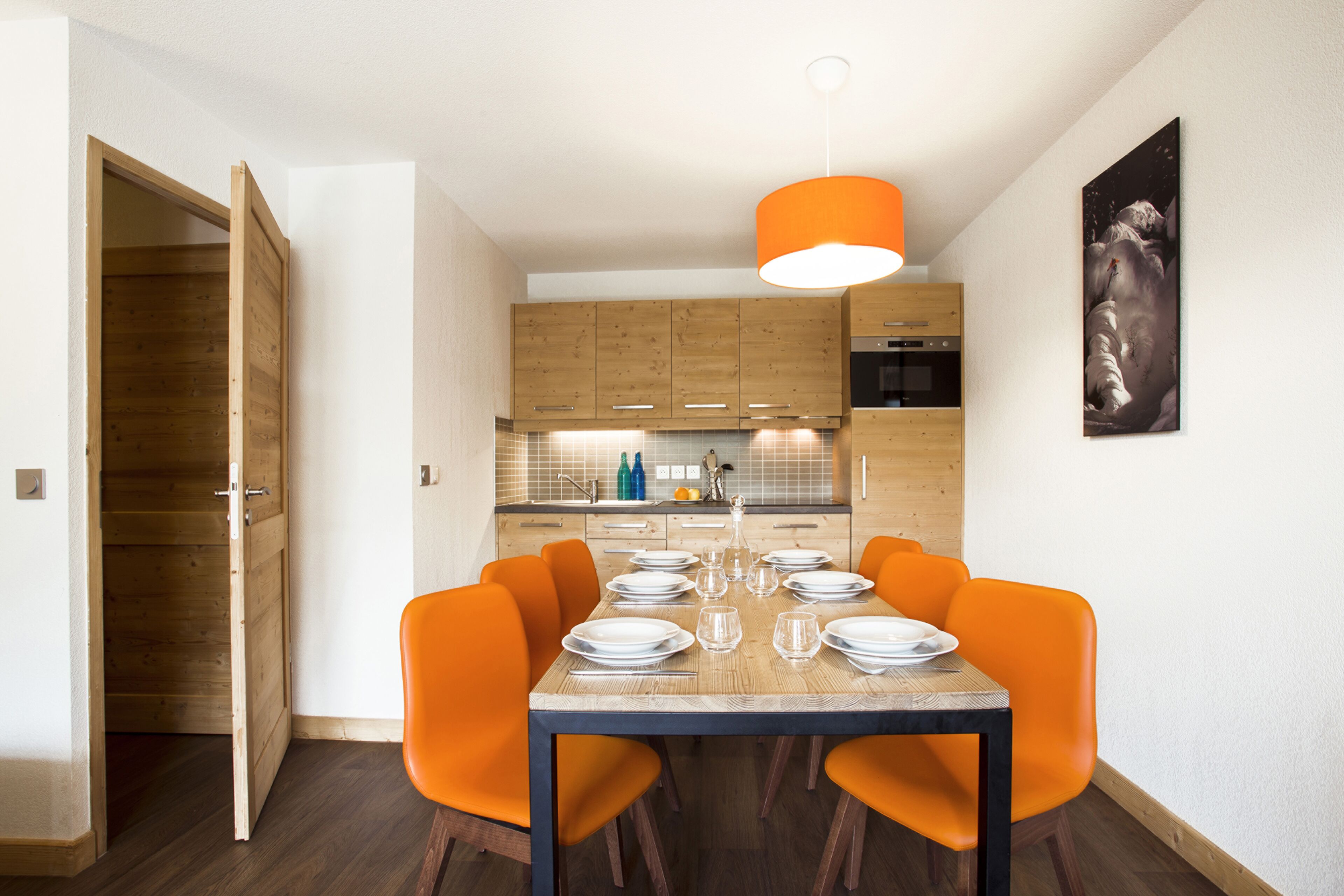 Een eetgedeelte met een houten tafel gedekt voor vier personen, omringd door oranje stoelen, in een moderne keuken met houten kasten en een hanglamp.