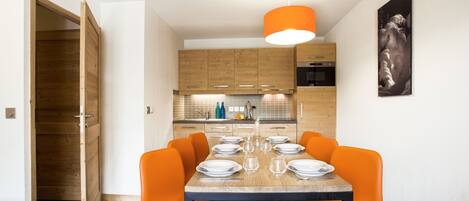 Een eetgedeelte met een houten tafel gedekt voor vier personen, omringd door oranje stoelen, in een moderne keuken met houten kasten en een hanglamp.