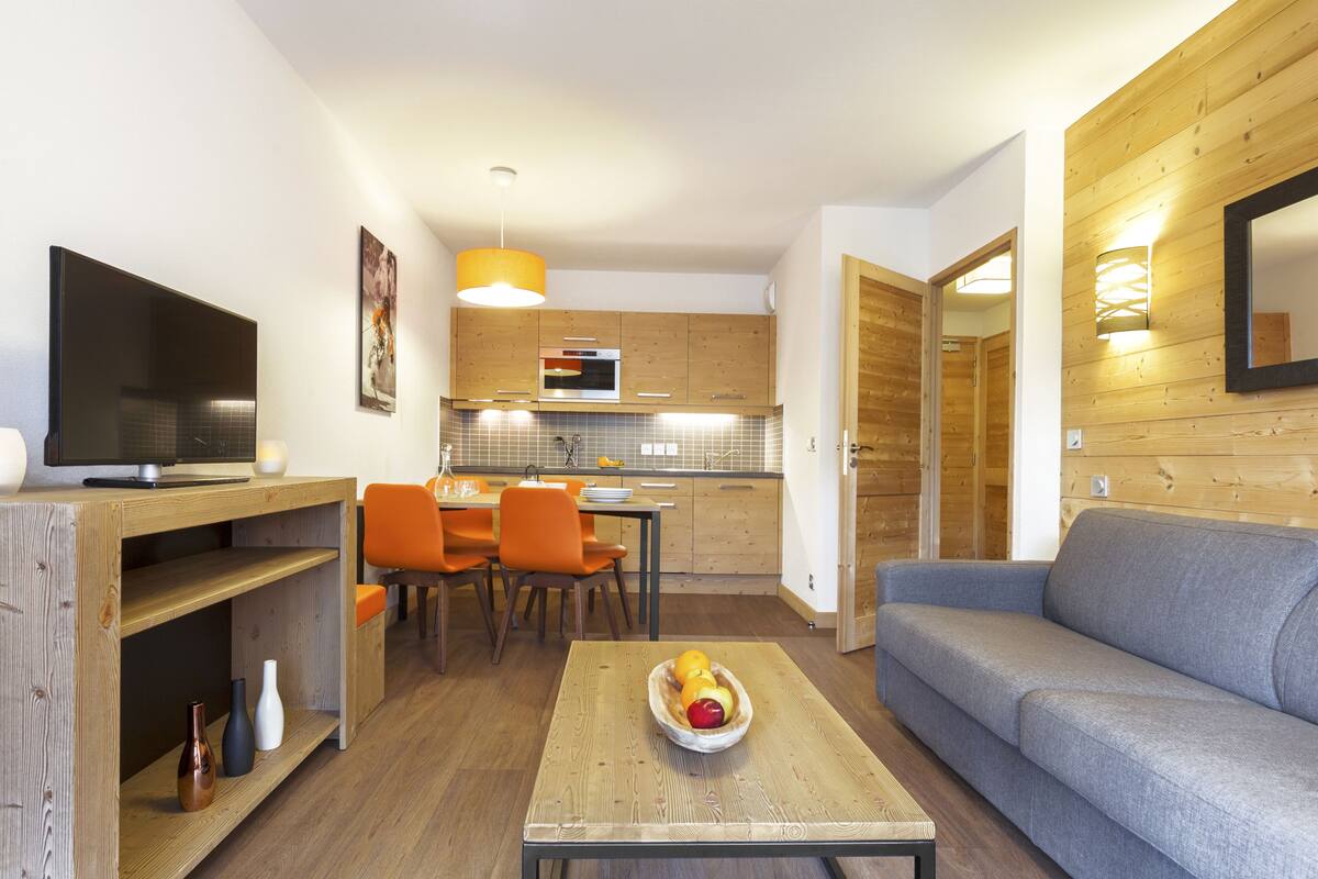 A modern living room with a grey sofa, wooden coffee table, and orange chairs. A flat-screen TV is on a wooden stand. The kitchen in the background has wooden cabinets and a microwave.