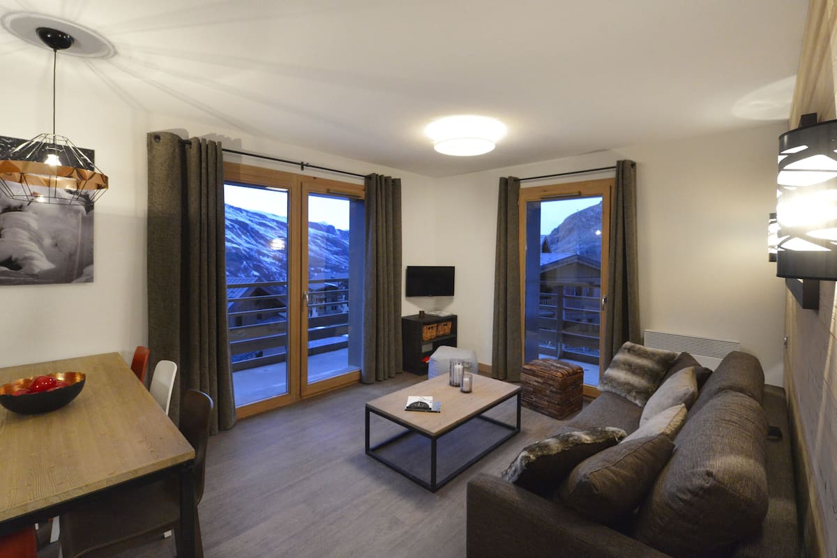 A modern living room with a wooden dining table, a sofa, a coffee table, and a TV on a stand. There are large windows with curtains, a painting on the wall, and a ceiling light.