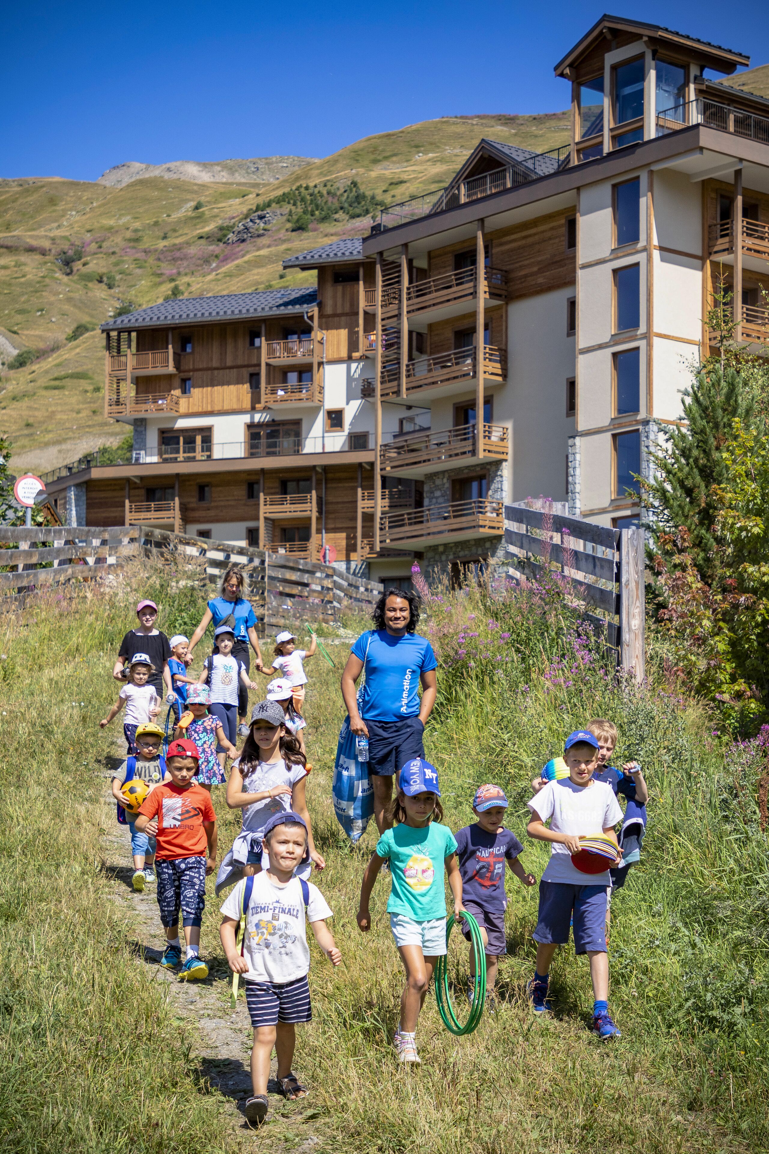A group of people walking on a path towards a multi-story building with balconies, surrounded by greenery and hills.