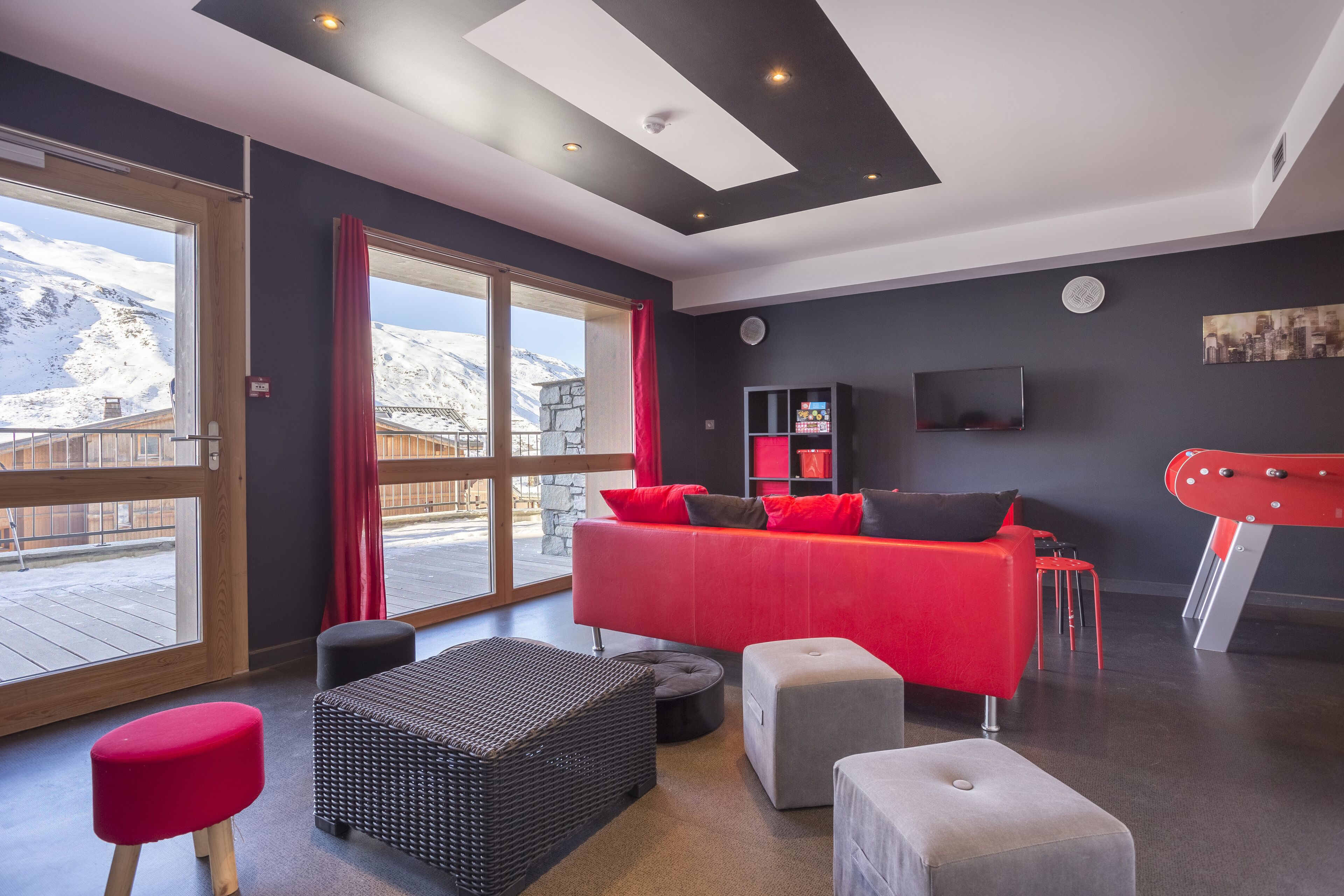 Uma sala de estar moderna com um sofá vermelho, pufes pretos e cinzentos, e uma janela grande com vista para montanhas nevadas.