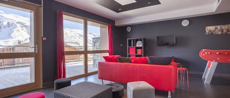 Uma sala de estar moderna com um sofá vermelho, pufes pretos e cinzentos, e uma janela grande com vista para montanhas nevadas.