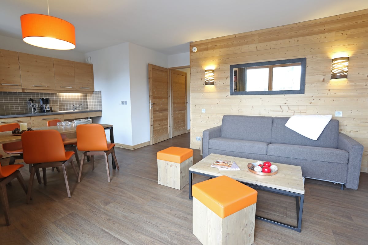 A modern living room with a grey sofa, orange chairs, a wooden coffee table, and a kitchen area with wooden cabinets and a bright orange pendant light.