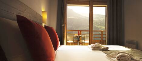 Habitación de hotel con cama, almohadas, una mesa con sillas y vistas a las montañas a través de un ventanal grande.