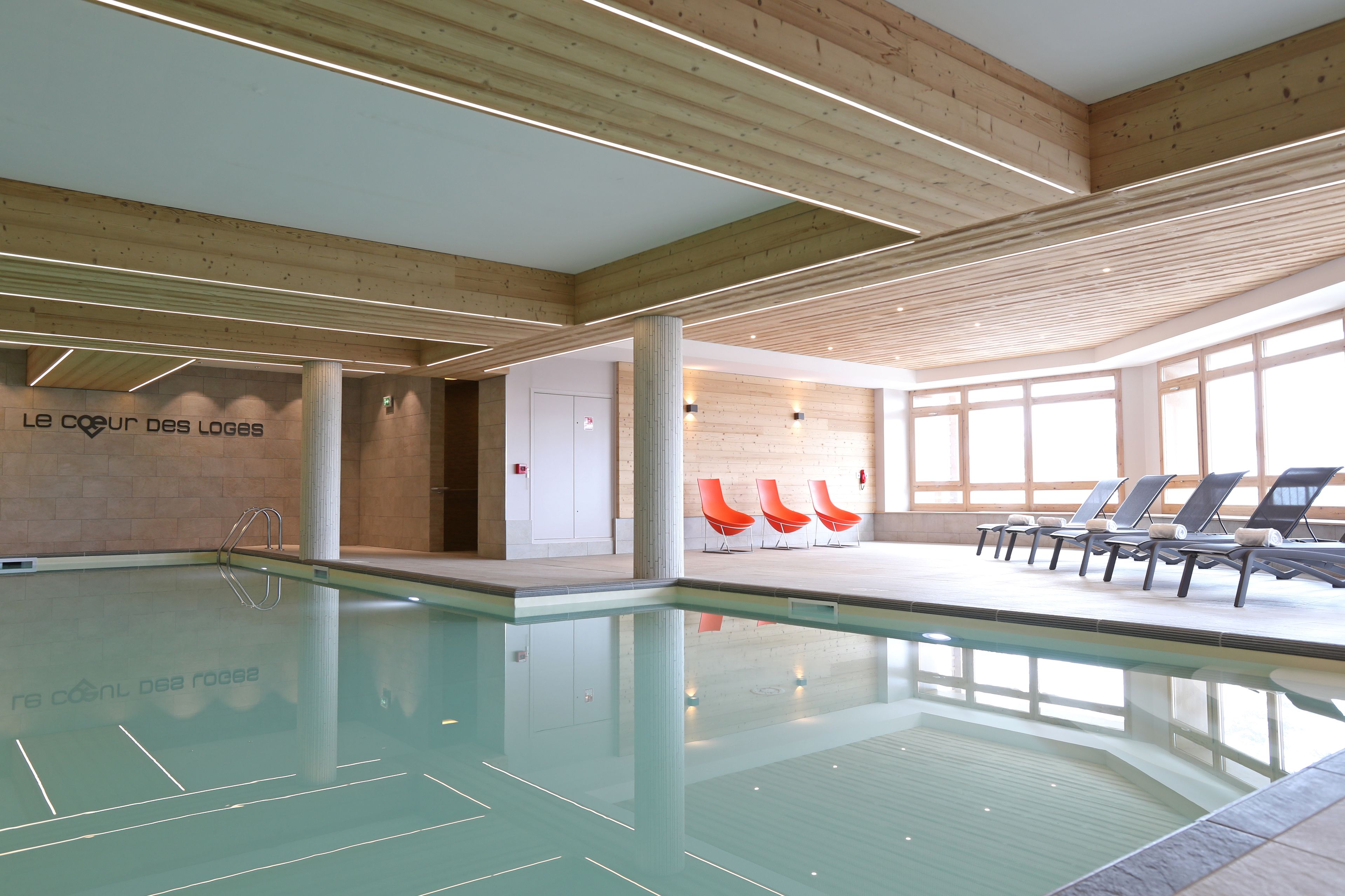An indoor swimming pool with wooden beams, a lounge area with orange chairs, and a wall with text.
