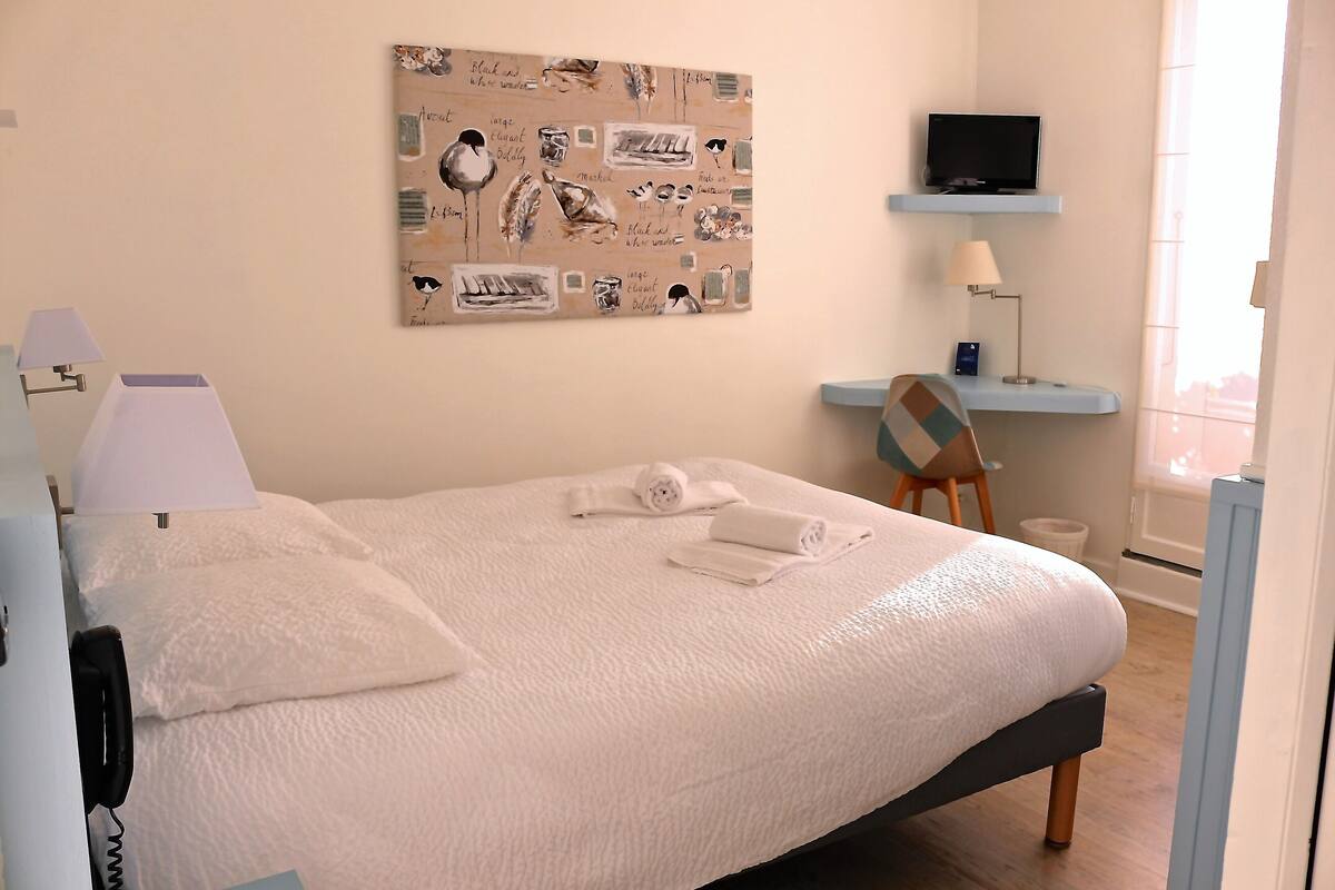 A neatly made bed with white linens, a mounted television above it, and a desk with a lamp to the right.