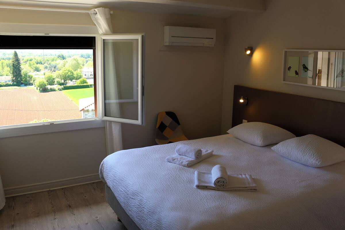 A hotel room with a large bed, a window with a view of a green landscape, an air conditioning unit, and a wall-mounted mirror.