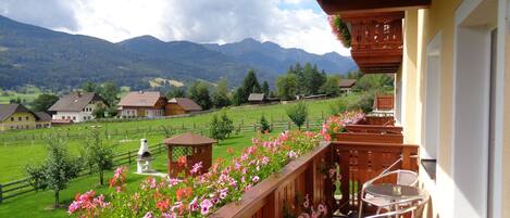 A balcony with a wooden railing, a small table, and chairs. In the background, there are houses, a fenced field, and mountains.