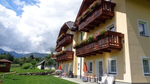 Un hotel con balcones de madera decorados con flores, un espacio exterior empedrado con sillas y mesas, y vistas a montañas al fondo.