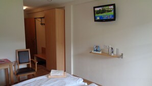 Habitación de hotel con cama, una silla, televisor montado en la pared y repisas con artículos de tocador.