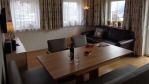 Una sala de estar moderna con una mesa de comedor de madera, dos sillones, un sofá y un televisor sobre un mueble bajo. Hay cortinas en las ventanas, una lámpara de pie y una planta en maceta en el alféizar.
