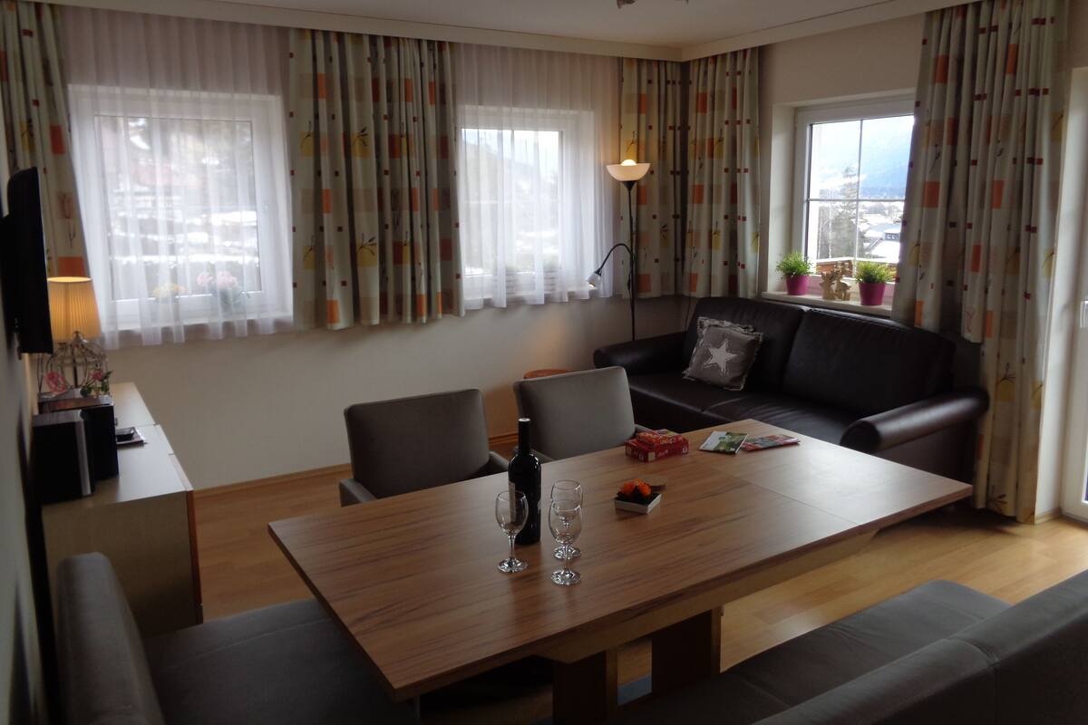 Una sala de estar moderna con una mesa de comedor de madera, dos sillones, un sofá y un televisor sobre un mueble bajo. Hay cortinas en las ventanas, una lámpara de pie y una planta en maceta en el alféizar.