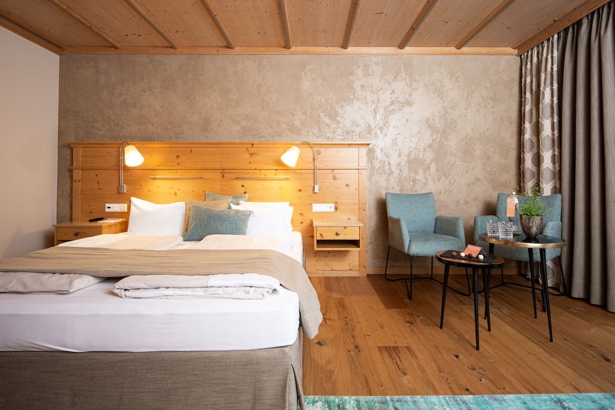 A hotel room with a wooden headboard, a bed with white linens, a small side table, a chair, a round table, and a potted plant.