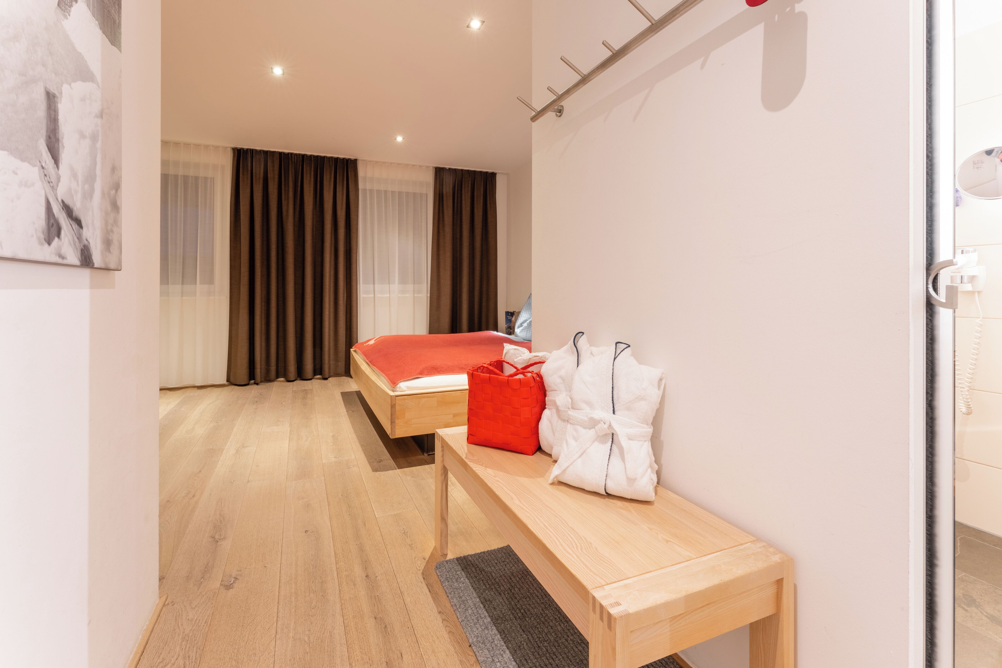 Ein Hotelzimmer mit Holzboden, einem Bett mit roter Decke, einer Bank mit weißen Handtüchern und einem Fenster mit Vorhängen.