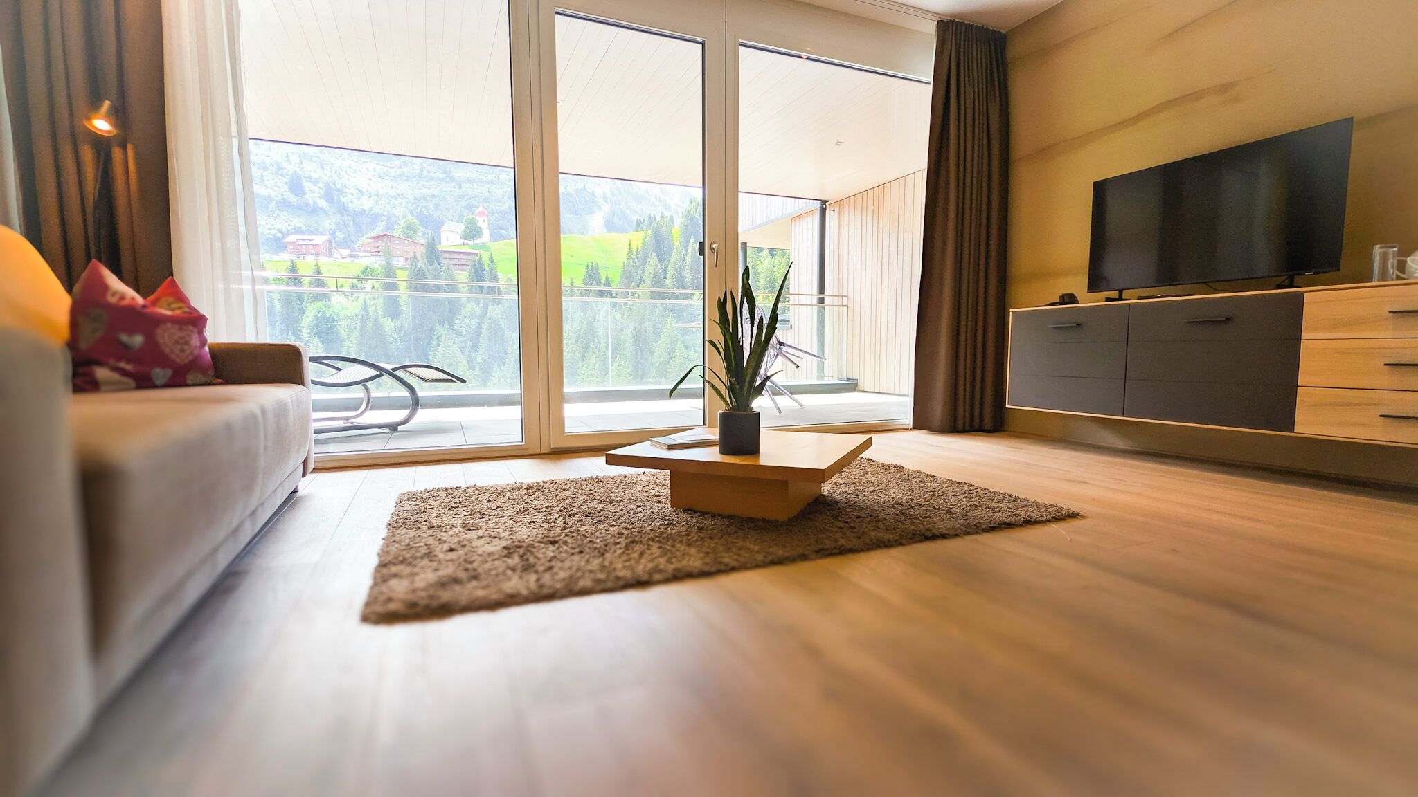 Ein modernes Wohnzimmer mit einer Couch, einem Couchtisch, einem Fernseher und einem großen Fenster mit Blick auf Grünflächen und Gebäude.