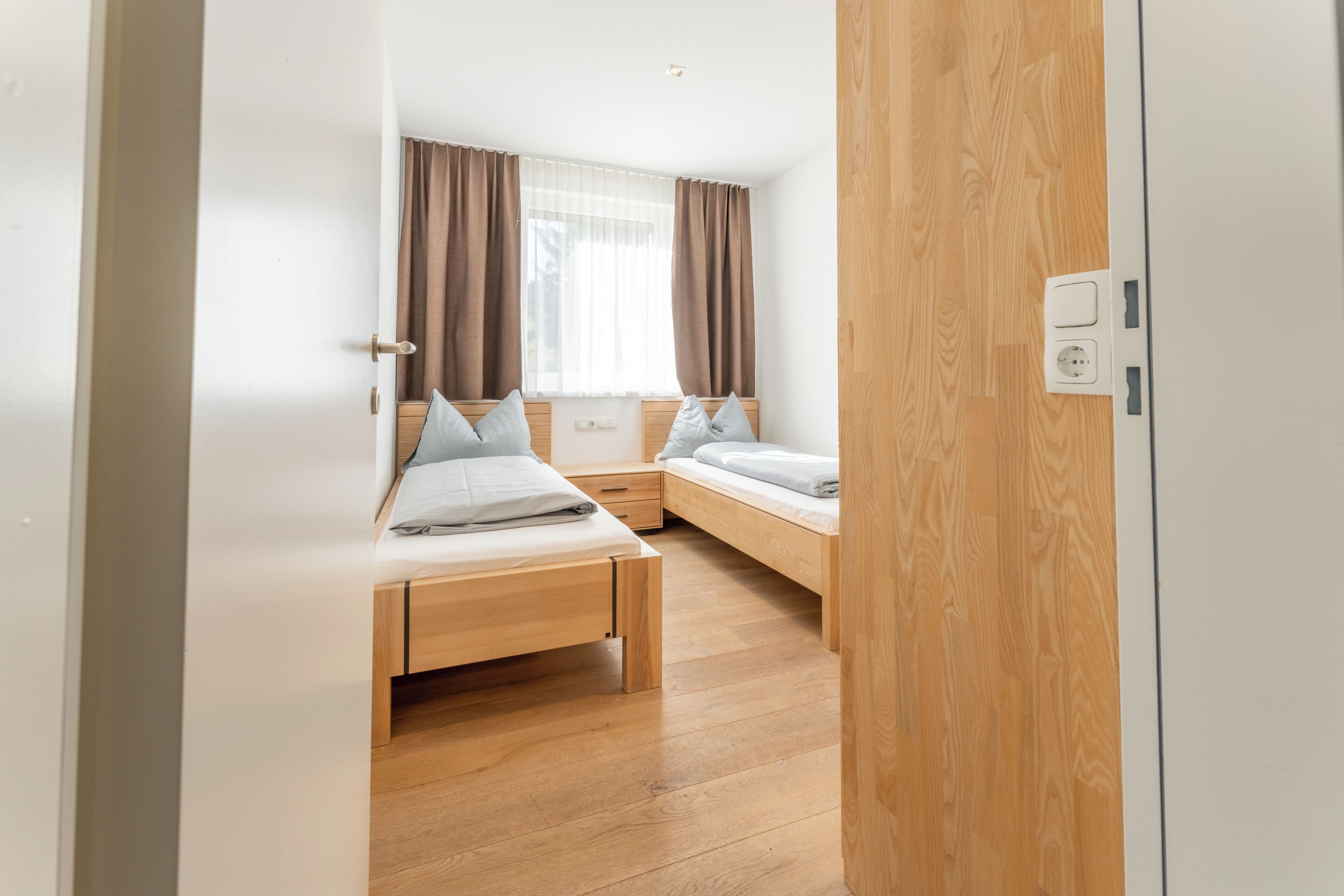 A modern bedroom with two single beds, a wooden floor, a window with curtains, and a white wardrobe.