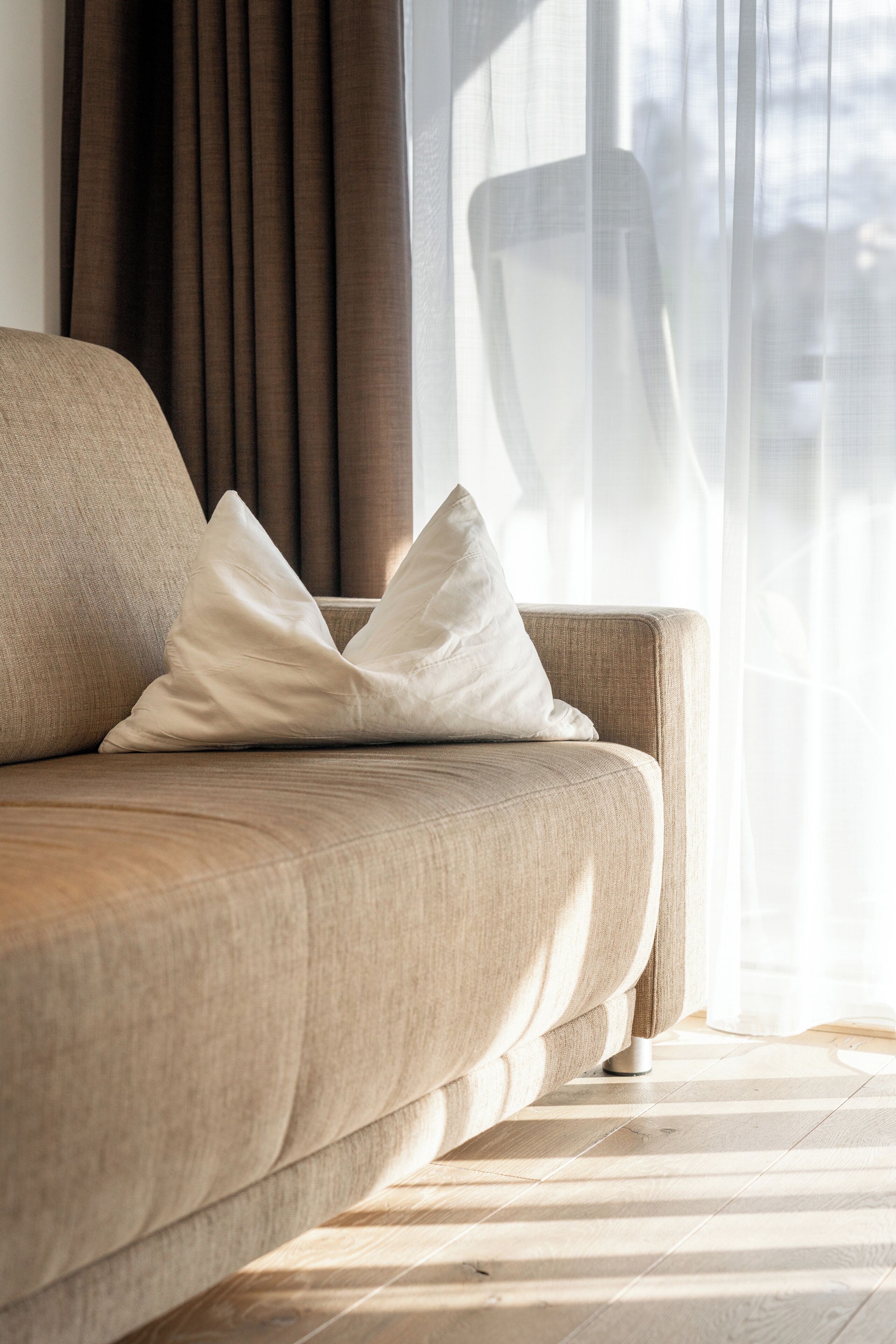 A beige sofa with a white pillow, positioned near a window with sheer curtains.