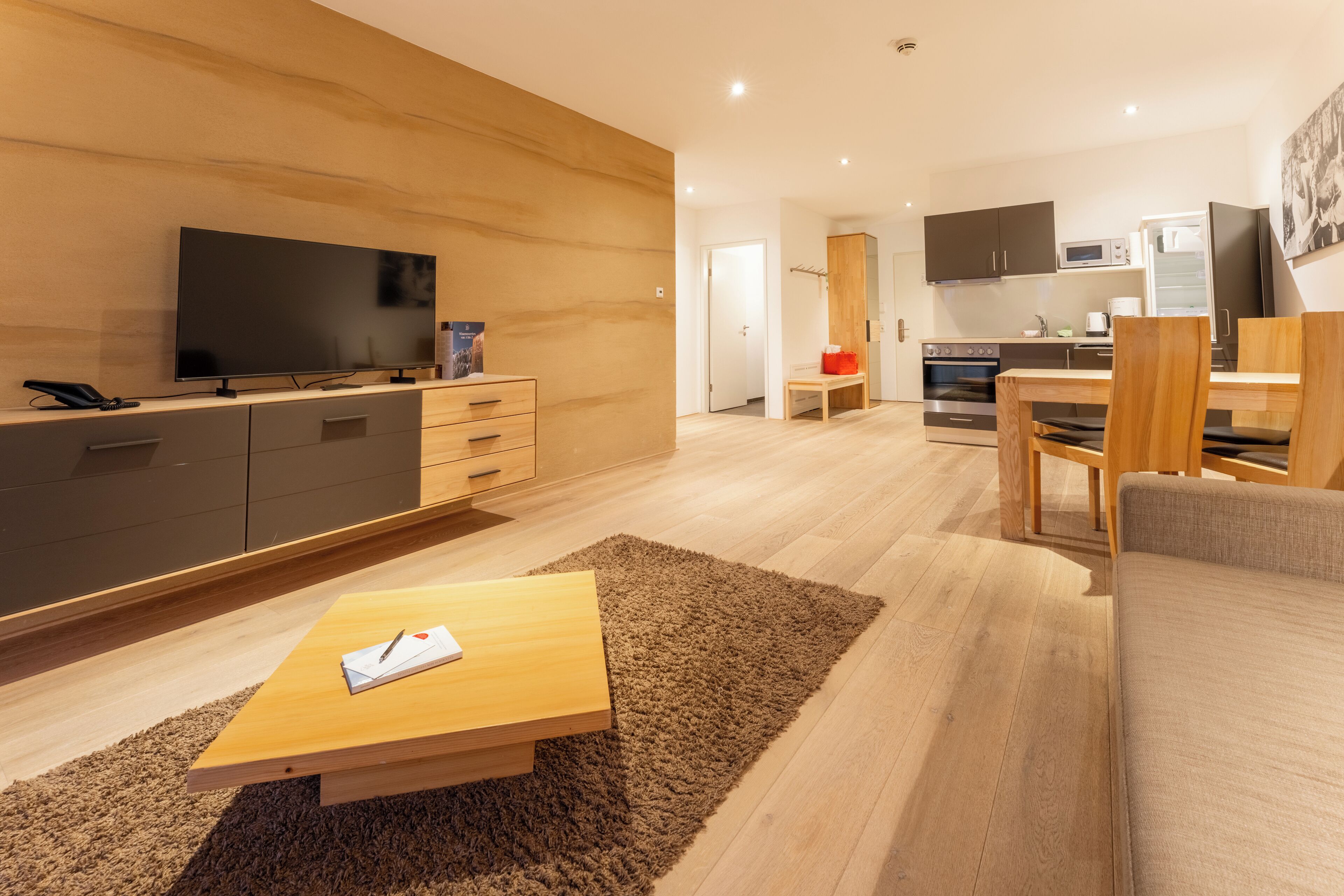 Een moderne woonkamer met een flatscreen televisie, houten meubels en een keuken op de achtergrond.