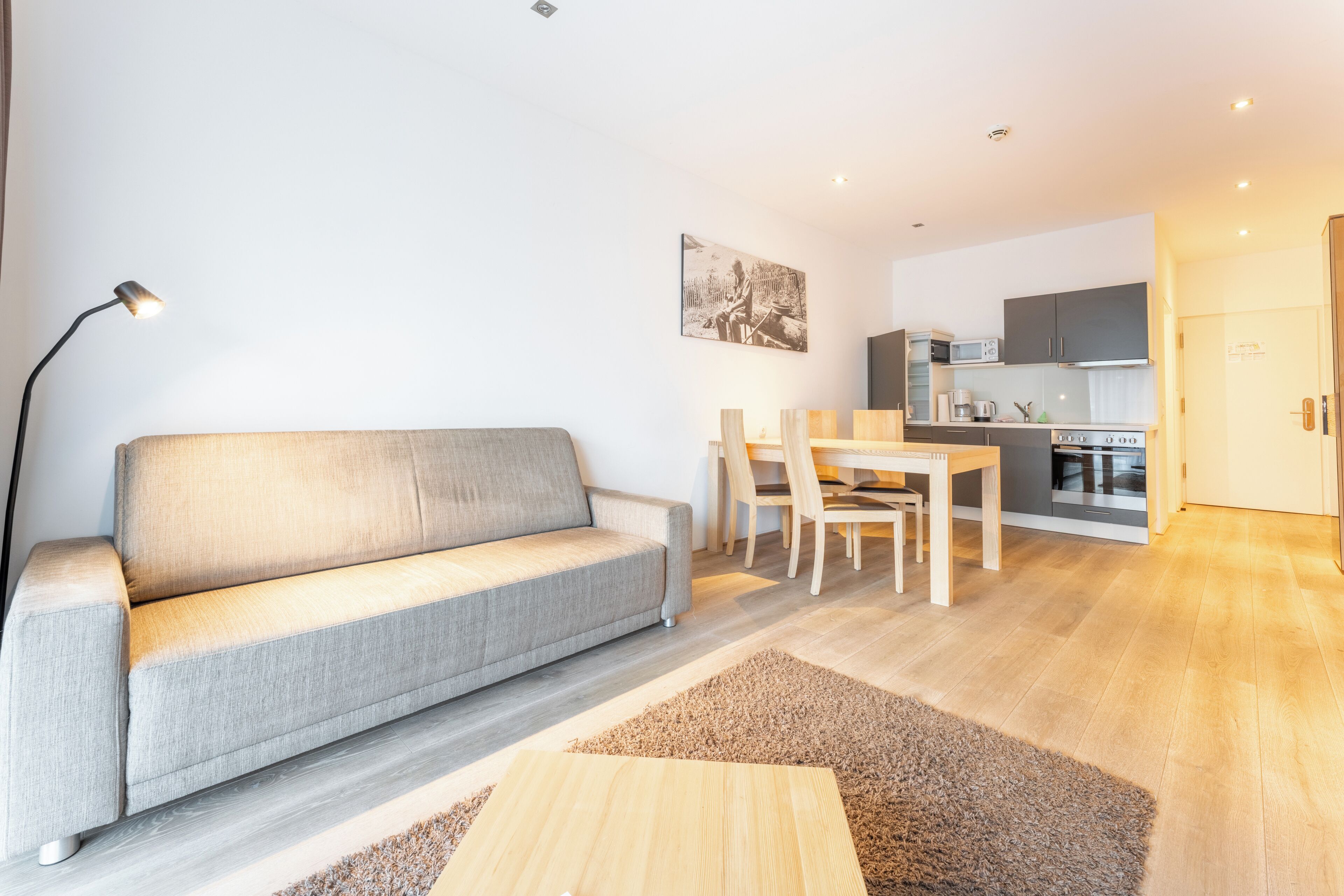 Ein modernes Wohnzimmer mit einem grauen Sofa, einem Holztisch und einem Essbereich mit Stühlen und einem Tisch. Im Hintergrund ist eine Küche mit Schränken und Geräten zu sehen.