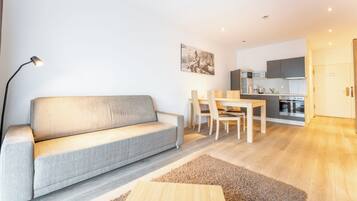 Ein modernes Wohnzimmer mit einem grauen Sofa, einem Holztisch und einem Essbereich mit Stühlen und einem Tisch. Im Hintergrund ist eine Küche mit Schränken und Geräten zu sehen.