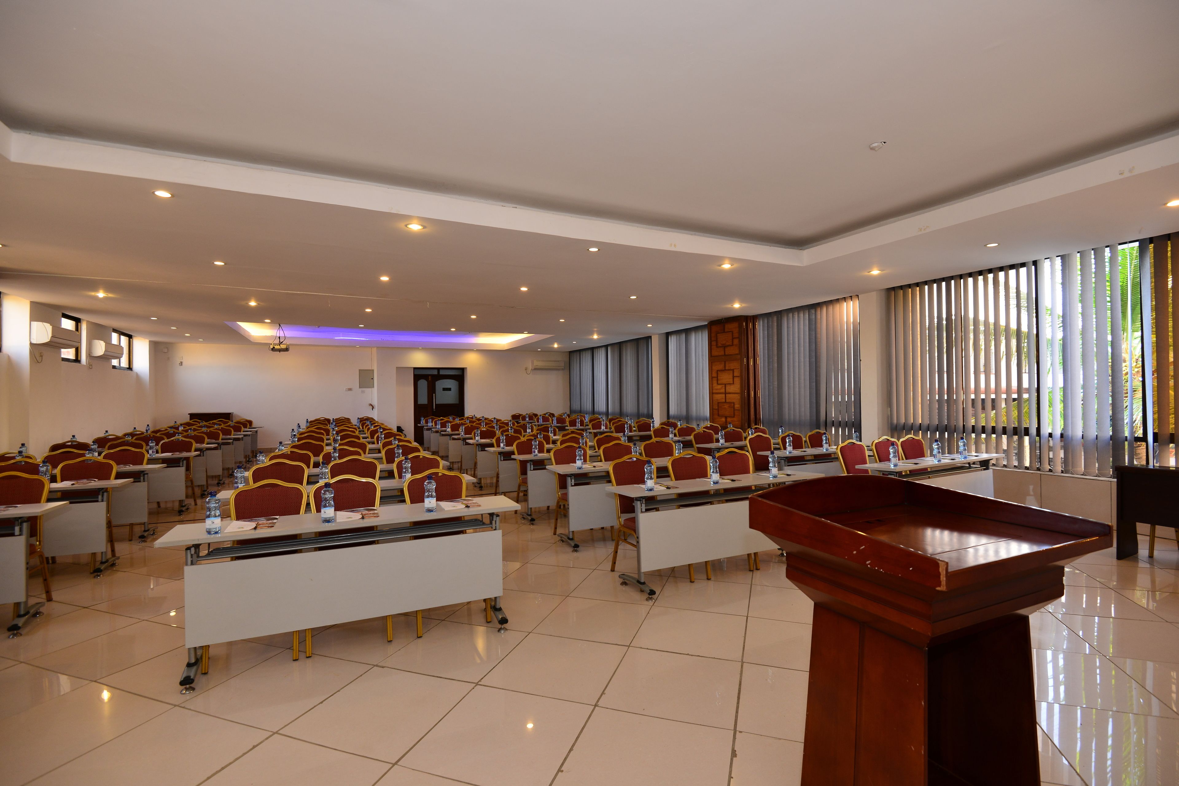 A conference room with rows of tables and chairs, a podium, and large windows with curtains.