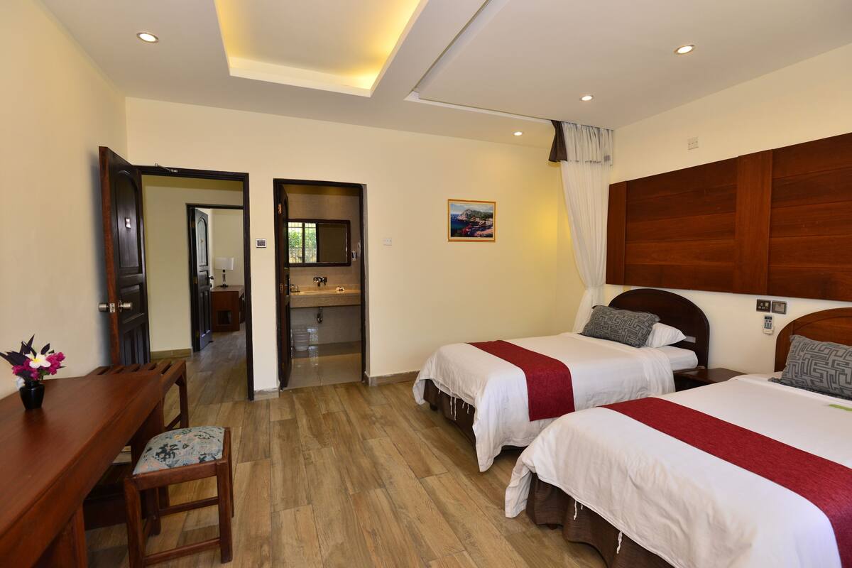 Habitación de hotel con dos camas, un escritorio de madera, una silla, una mesita con un florero y un baño visible a través de una puerta abierta.