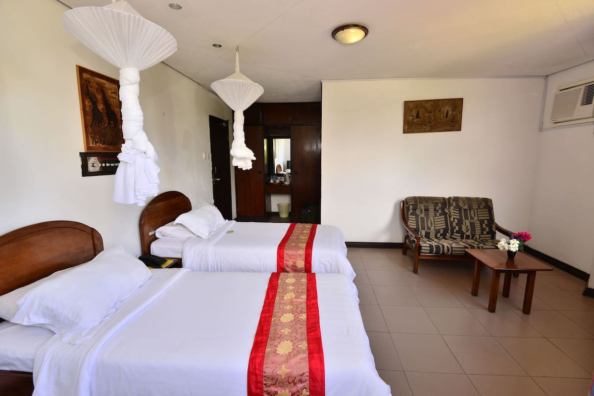 Habitación de hotel con dos camas, cabecero de madera, un sillón estampado, una mesita con un jarrón y una lámpara colgante.