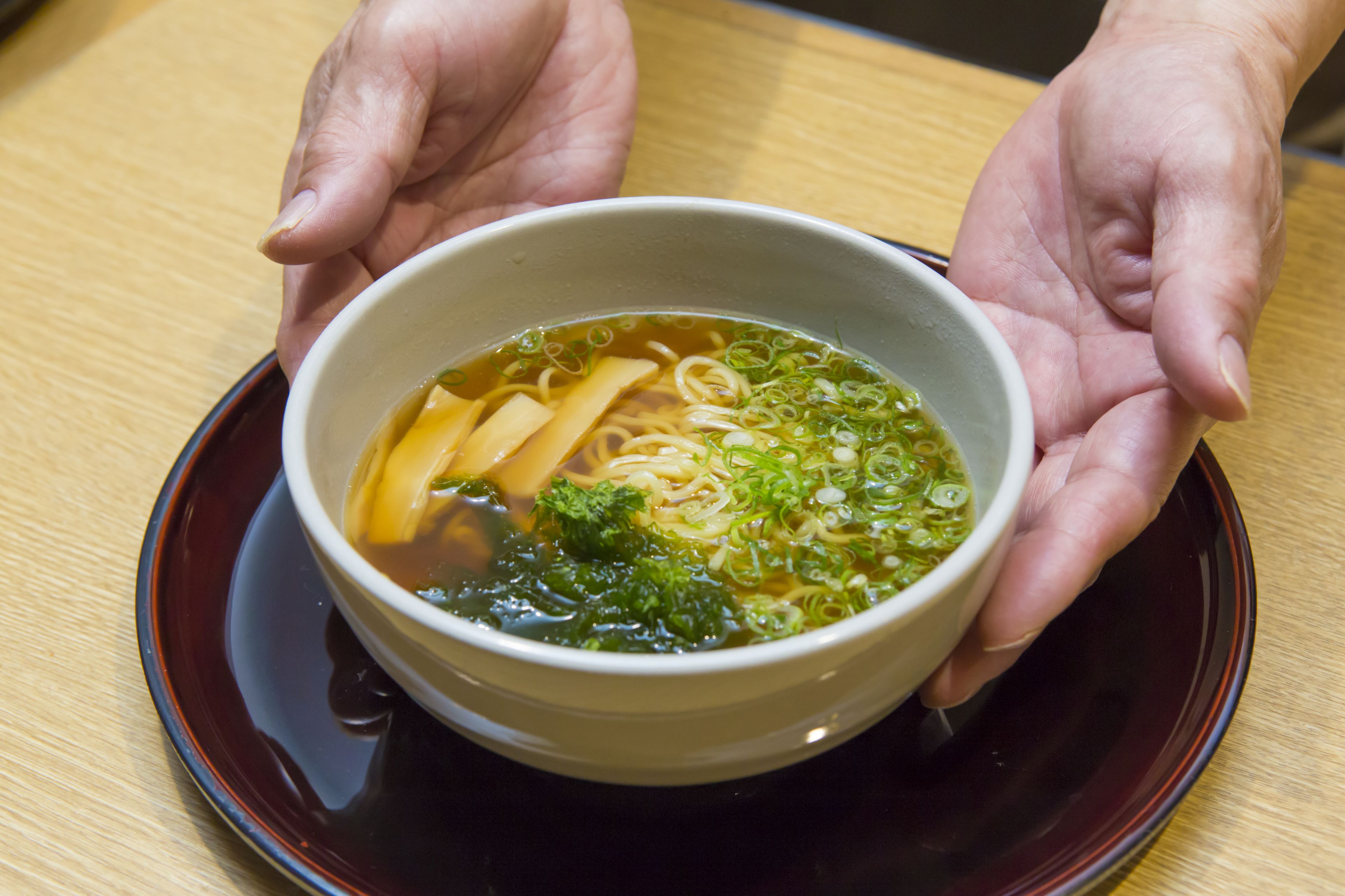A bowl of ramen held by hands over a dark saucer on a wooden table.