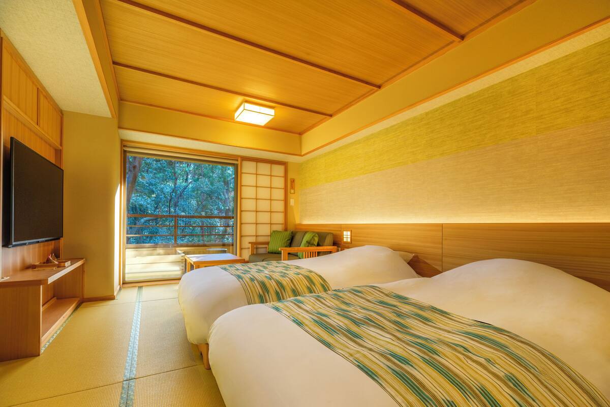 A traditional Japanese-style room with tatami flooring, a bed with a striped blanket, a television, and a sliding door leading to a balcony with a view of trees.