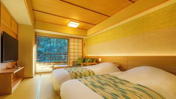 Ein traditioneller japanischer Raum mit Tatami-Boden, einem Bett mit Streifenmusterdecke, einem Fernseher und einer Schiebetür, die auf einen Balkon mit Blick auf Bäume führt.