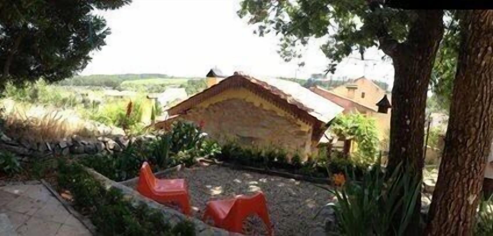 A rustic stone building with a sloped roof, surrounded by greenery and a paved area with two red plastic chairs.