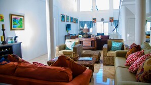 Ein geräumiges Wohnzimmer mit Rattanmöbeln, einer Couch und einem Couchtisch. An den Wänden befinden sich mehrere gerahmte Kunstwerke und ein Fenster sorgt für natürliches Licht.