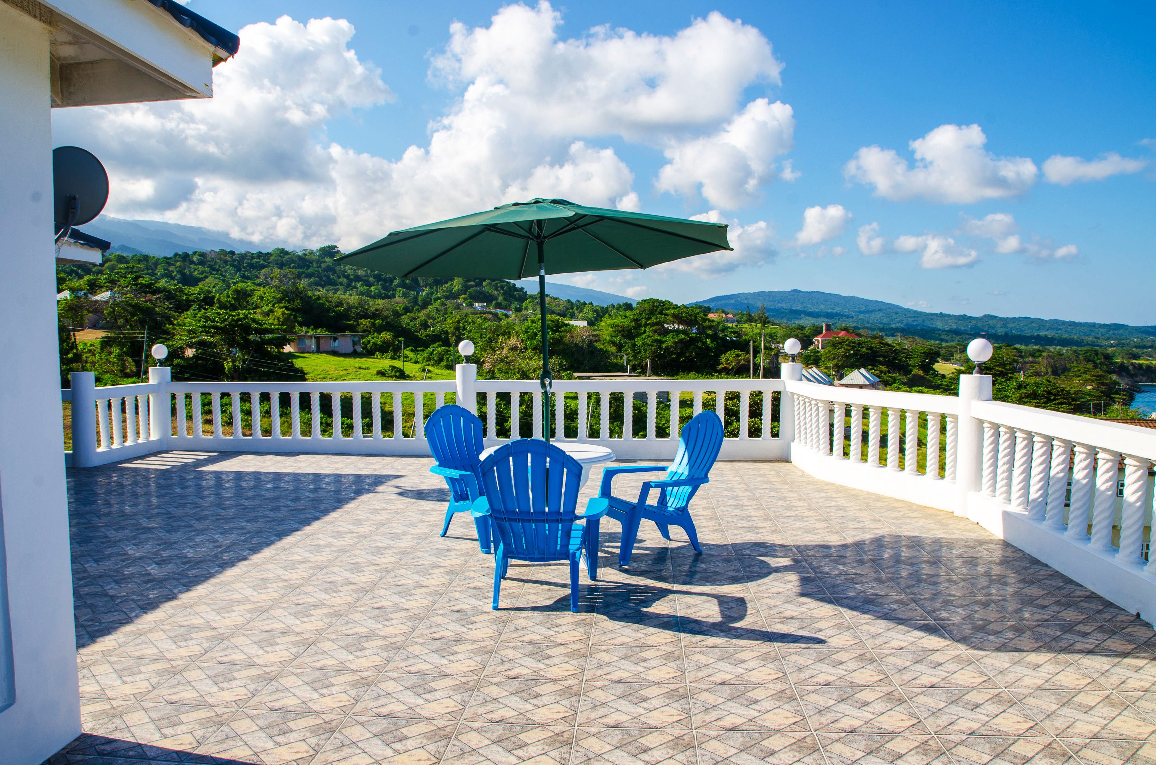 Eine Terrasse mit blauen Stühlen und einem Tisch, einem grünen Sonnenschirm und einem weißen Geländer.