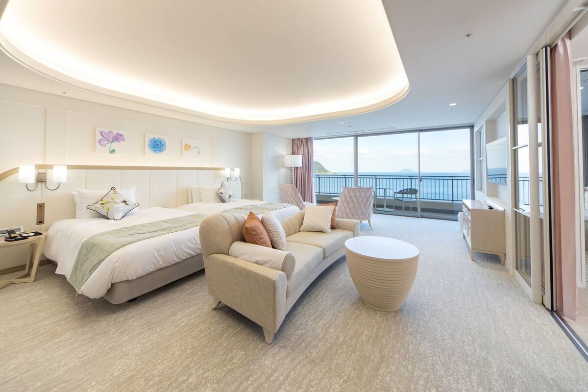 Ein modernes Hotelzimmer mit einem großen Bett, einer Couch und einem runden Couchtisch. Ein Balkon bietet Blick auf das Meer.