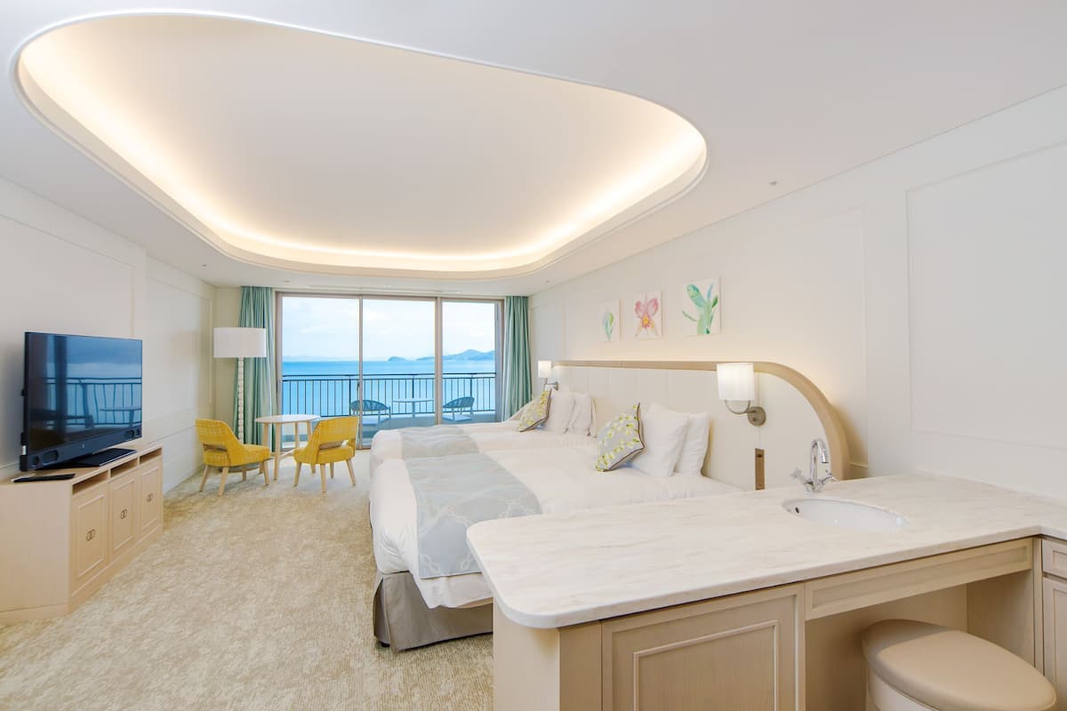 Ein Hotelzimmer mit einem großen Bett, einem Fernseher, einem Schreibtisch mit Stühlen, einem Badezimmer mit Waschbecken und einem Balkon mit Meerblick.