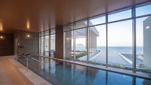 A modern indoor infinity pool with clear glass walls, a wooden ceiling, and a view of the sea. - Karatsu Seaside Hotel (Karatsu)
