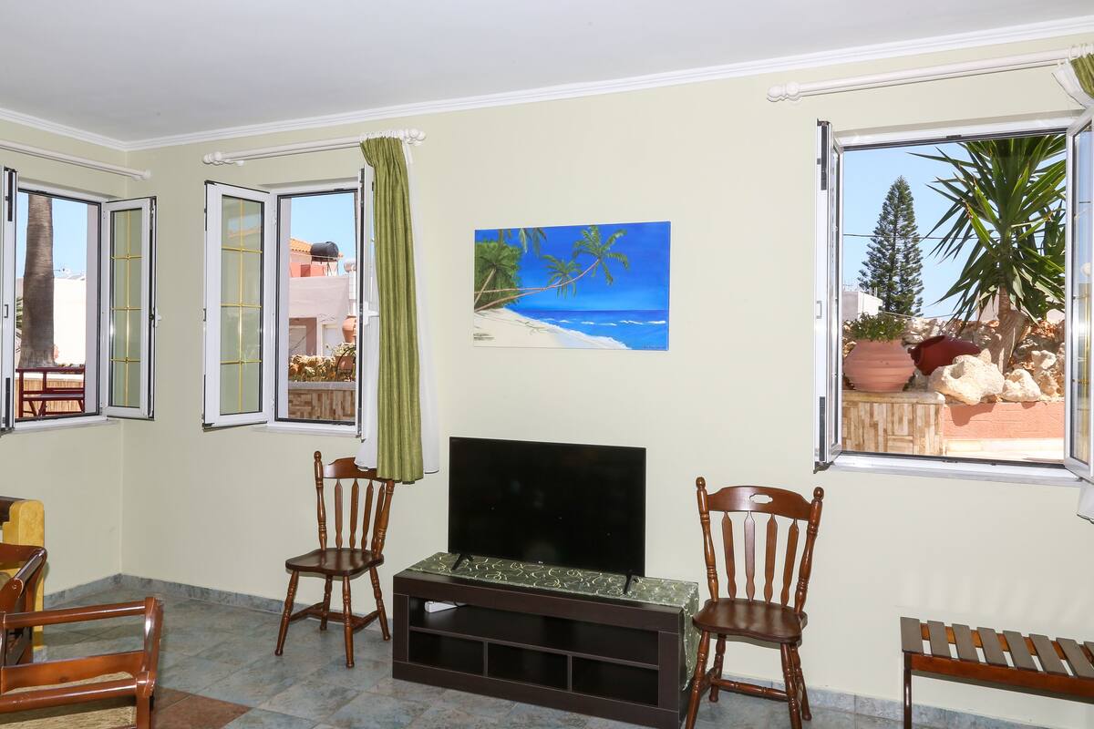 Ein Wohnzimmer mit Flachbildfernseher, zwei Holzstühlen, einer Bank und einem Gemälde mit Strandmotiv.